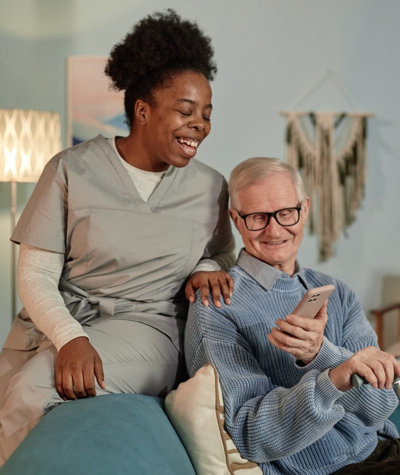 Smiling Black Female Caregiver Helping Elderly Man Using Phone Indoors